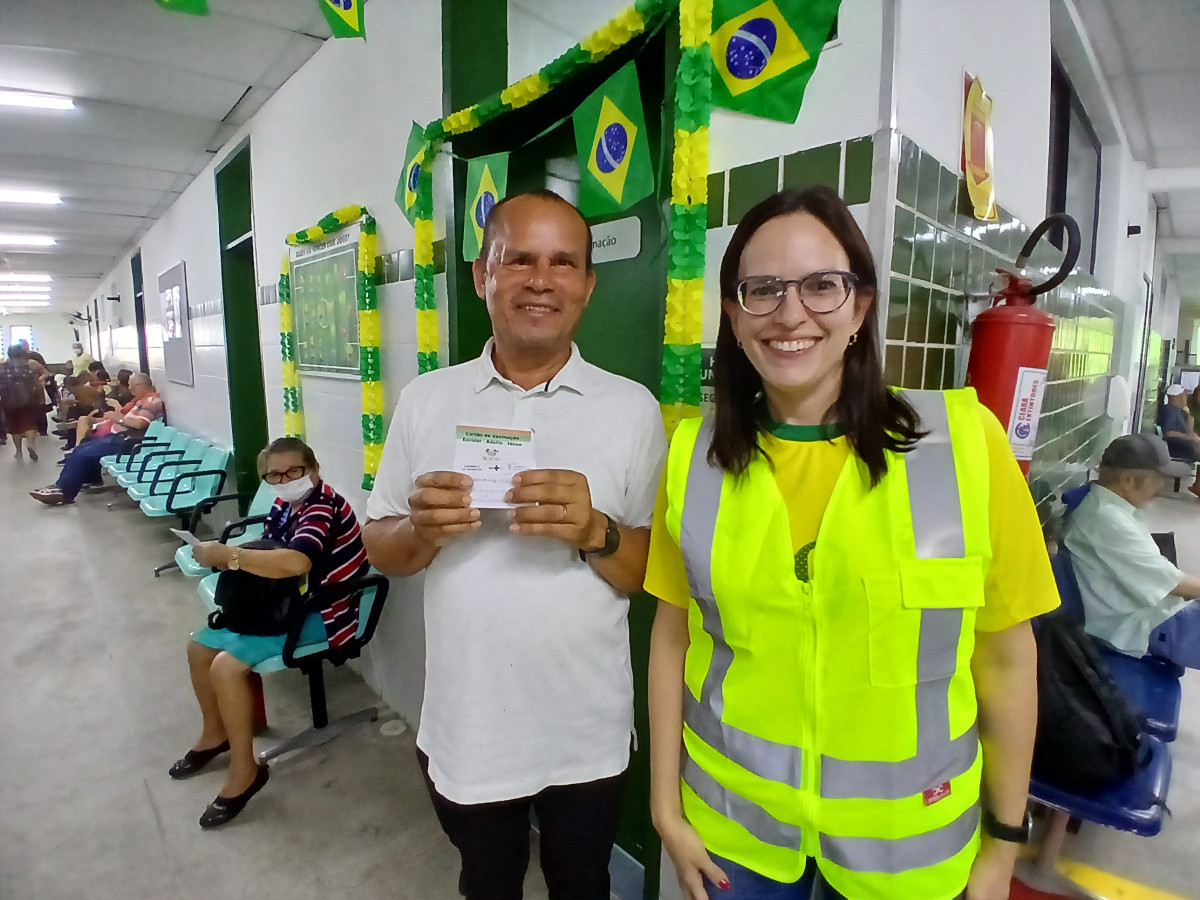 Paciente mostra cartão de vacina no CIDH ao lado de enfermeira vestida com colete de fiscalização