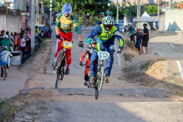 Fotografia com dois ciclistas em destaque. Os dois usam capacete e roupas coloridas de competição, e estão pedalando em uma rua. Há pessoas nas calçadas acompanhando a competição.